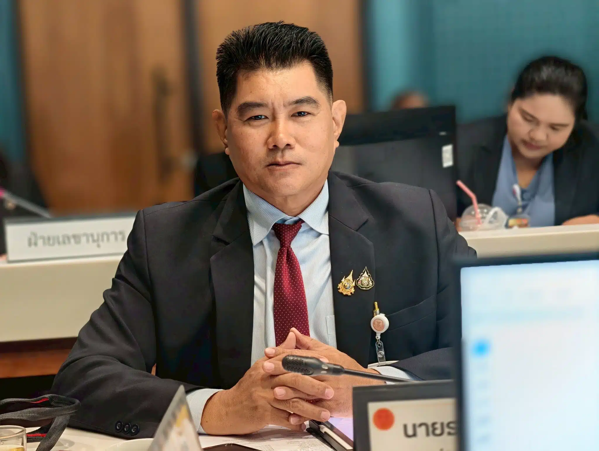 ภาพธนกร ถาวรชินโชติ กำลังนั่งอยู่ในห้องประชุม แสดงท่าทีจริงจังในชุดสูท