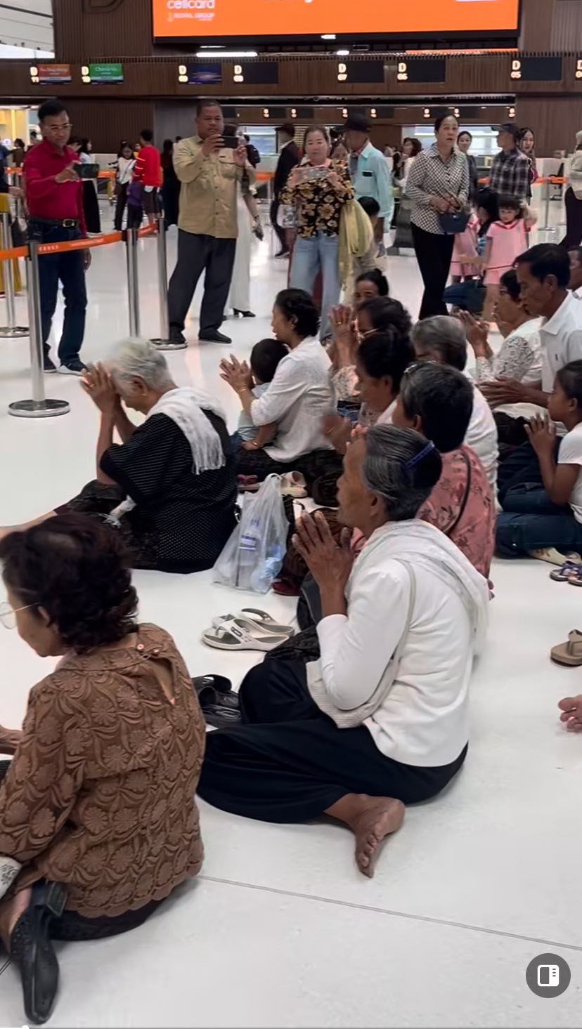 กลุ่มคนชาวกัมพูชาในชุดขาวกำลังนั่งพับเพียบไหว้พระพุทธรูปสีทองในอาคารผู้โดยสาร