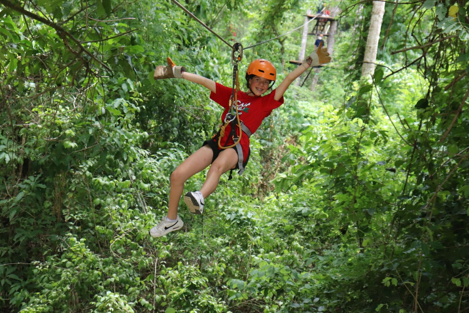 A kid wearing red t-shirt smiling while zip lining through the forest during summer camp at iCamp Thailand.