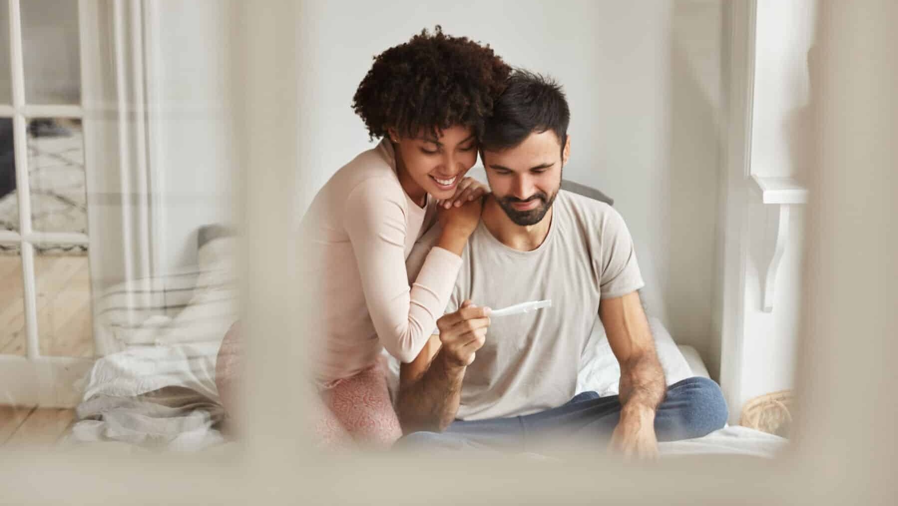Happy mixed race future parents look joyfully at pregnancy test, rejoice positive news about pregnancy, sit together on bed against domestic interior. Newlywed couple wait for new member in family after IVF in Thailand