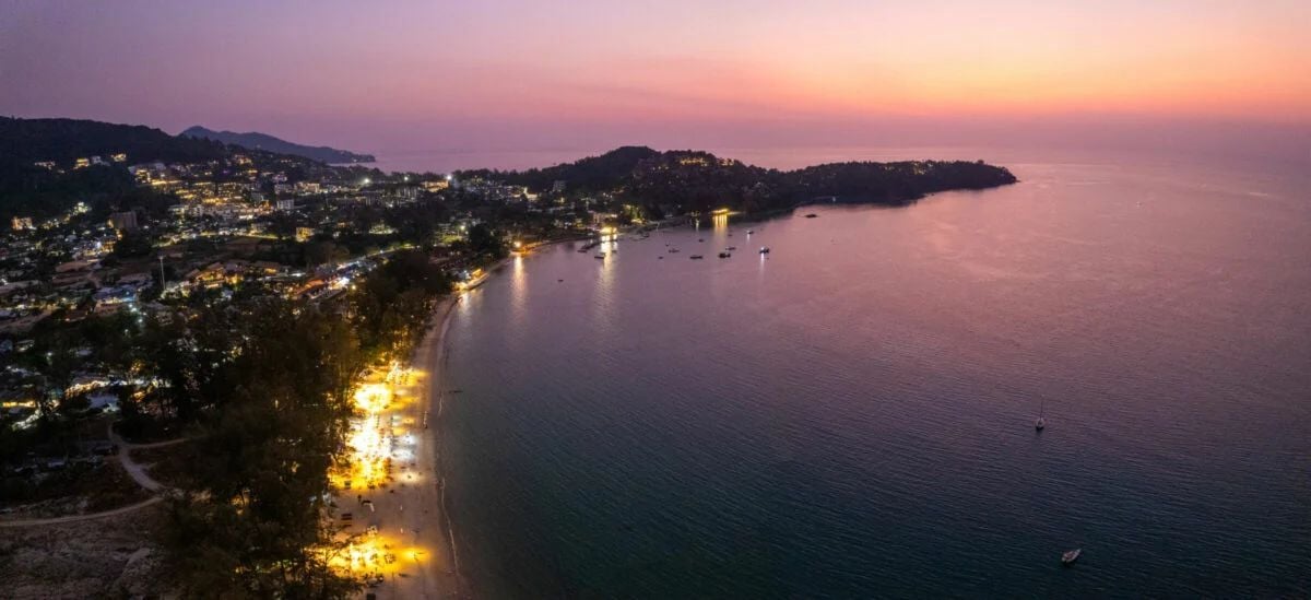 Aerial view of Bang Tao beach, one of the best places to live in Phuket, at sunset with glowing city lights and calm sea.