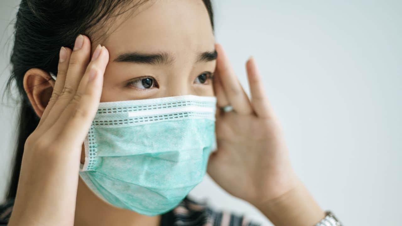 Woman wearing a face mask holding her head in pain, showing flu-like symptoms